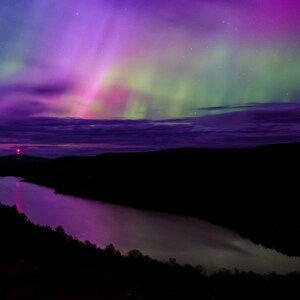 May include: A vibrant aurora borealis display in shades of purple, green, and pink over a dark, still lake. The aurora is reflected in the water, creating a mesmerizing scene.