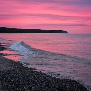 May include: A pink and purple sunset over a calm lake with a sandy beach and a small wave breaking on the shore.