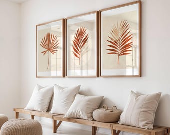 May include: Three framed botanical prints with brown wooden frames. Each print features a different stylized illustration of a reddish-orange palm leaf against a cream-colored background. The prints are displayed above a wooden bench with white pillows.