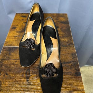 Op de afbeelding: Een paar zwarte suède pumps met een lage hak. Elke schoen heeft een decoratieve zwarte stoffen bloem op de wreef. De binnenkant van de schoenen is bruin en de zolen zijn zwart. De schoenen worden op een houten oppervlak gepresenteerd.