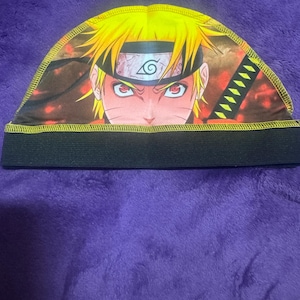 May include: A black and yellow skullcap with a colorful anime character print. The character has spiky yellow hair, red eyes, and a headband with a swirl symbol.