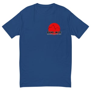 May include: A blue t-shirt featuring a red sun graphic with a mountain range silhouette and the text "DYNO-MITE". Below the text is the phrase "DYNAMIC CLIMBING GYM MACHO PICCHU". The shirt is made of a soft material.
