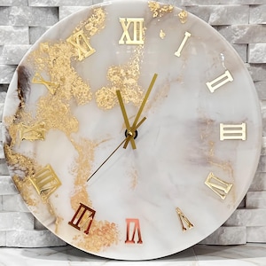 May include: A round wall clock with a white and gold marble design. The clock face features gold Roman numerals and gold leaf accents. The clock hands are gold. The clock is displayed on a white surface against a white brick wall.