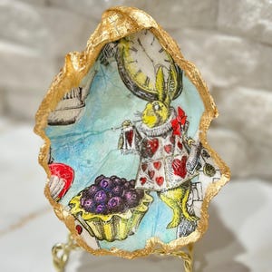 May include: A gold-leafed seashell with a whimsical Alice in Wonderland design featuring the White Rabbit holding a pocket watch and a plate of tarts. The seashell is on a gold stand.