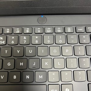 May include: Close-up of a black laptop keyboard with gray keys. The keyboard features function keys, number keys, and letter keys. The power button is illuminated with a blue light. The keys are labeled with symbols and characters.