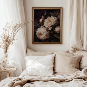 May include: A framed floral painting featuring large, white peonies with yellow centers and green leaves against a dark background. The painting is in a gold frame and hangs above a bed with beige bedding and pillows.