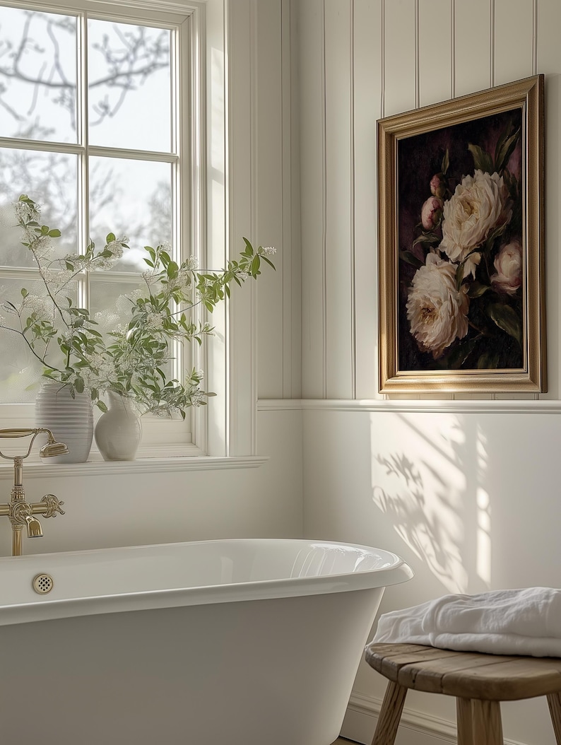 May include: A bathroom scene featuring a white clawfoot bathtub, a framed floral painting, and a wooden stool with a folded white towel. The room has a window with a view and vases with greenery.