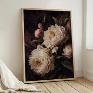 May include: A framed floral print featuring large, creamy white peonies with hints of pink against a dark, moody background. The painting style is reminiscent of classic oil paintings, with visible brushstrokes and a soft, romantic feel. The frame is a light wood.