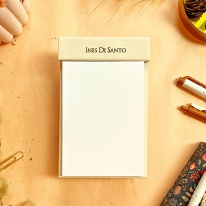 May include: A beige notepad with a white paper insert and a beige top that says "Ines Di Santo". The notepad is on a beige surface with gold glitter.