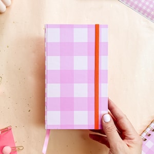 May include: A pink and white gingham pattern notebook with an orange elastic band closure and a pink ribbon bookmark. The notebook is held by a person's hand. Other stationery items are in the background.
