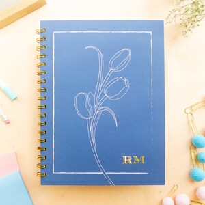 May include: A blue spiral notebook with a white outline of a tulip bouquet and the initials "RM" in gold lettering.