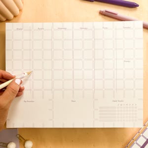 May include: A weekly planner with a grid layout, featuring sections for each day of the week, top priorities, notes, and a habit tracker. The planner is light beige with a subtle geometric pattern and includes a pencil.