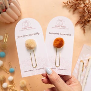 以下が含まれることがあります： 白いカードに「Paper Rosie」と「pompom paperclip」の文字が書かれた、金色のポンポン付きペーパークリップが2つ。1つのペーパークリップには薄い茶色のポンポン、もう1つのペーパークリップにはオレンジ色のポンポンが付いている。