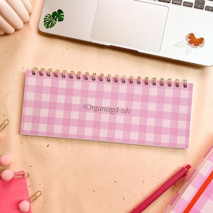 May include: A pink and white gingham notepad with gold spiral binding. The notepad features the text "Organized-ish". A pink pen and other stationery items are also visible.