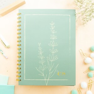 May include: A green spiral bound notebook with a gold wire binding. The cover features a white line drawing of lavender flowers and the initials "CS" in gold.