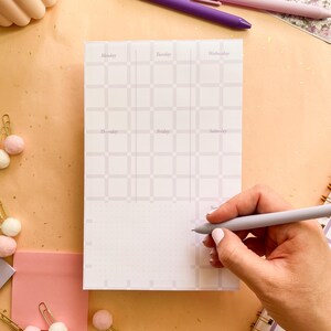 May include: A white and light purple weekly planner pad with a grid pattern and days of the week labels. A hand holds a light purple pen, poised to write on the pad. The pad is on a light orange surface.