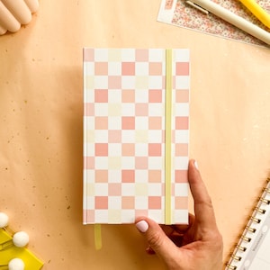 Könnte beinhalten: Ein Notizbuch mit einem rosa, pfirsichfarbenen und cremefarbenen Schachbrettmuster. Es hat einen gelben Gummibandverschluss und ein passendes Schleifenlesezeichen. Das Notizbuch wird von einer Hand mit hellrosa Maniküre gehalten.