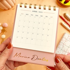 May include: A pink desk calendar with a gold spiral binding. The calendar is for April 2025 and has the name "Marina Dantas" printed on the bottom.