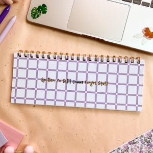 Op de afbeelding: Een spiraalgebonden notitieblok met een wit en lavendel geruit patroon. Het notitieblok heeft gouden letters met de tekst "Spoiler: I'm Still Gonna Forget Stuff". Een laptop en pennen staan op de achtergrond.
