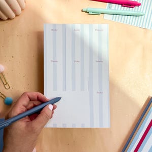 Puede incluir: Un planificador semanal con rayas verticales de color azul claro y los días de la semana impresos en una fuente púrpura suave. Una mano sostiene un bolígrafo azul, lista para escribir. El planificador está sobre una superficie marrón claro.
