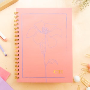May include: A spiral-bound notebook with a coral cover featuring a light blue floral line drawing and a gold foil monogram "CH". The notebook has a gold spiral binding and is surrounded by stationery items.