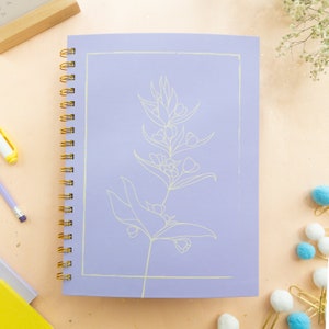 May include: A lavender notebook with a gold spiral binding. The cover features a white line drawing of a flower with a simple design.