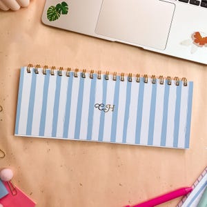May include: A notebook with a blue and white striped design, bound with a gold spiral. The initials "CH" are in gold script on the cover. The notebook is on a peach-coloured surface, near a laptop.