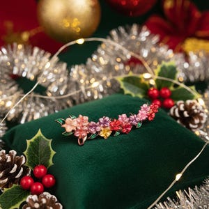 May include: A floral brooch with pink, red, and yellow flowers, set against a green velvet cushion. The brooch is accented with green leaves and a gold-tone metal. Christmas ornaments and tinsel are in the background.