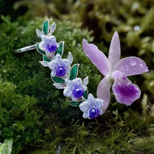 May include: A silver ear cuff featuring a vertical arrangement of four white and purple orchid-shaped flowers with green leaves and small clear stones. The jewelry piece is designed to follow the curve of the ear and displays intricate enamel detailing.