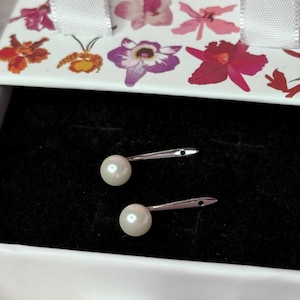 May include: Two pearl earrings with silver posts are displayed in a white box with a black interior. The box lid is decorated with colorful floral designs. The earrings are round and white.