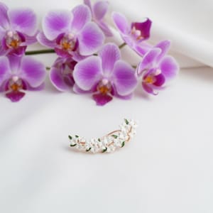 May include: A gold-toned floral hair clip with white flowers and green leaves. The clip is curved and rests on a white surface, with purple orchids in the background.