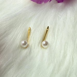 May include: A pair of gold-colored earrings with a pearl accent. The earrings have a long, slender design with a small, round pearl at the bottom. The background is a soft, white, furry texture, with a purple flower in the upper right corner.