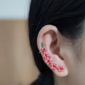 May include: Ear cuff featuring a floral design. The ear cuff is adorned with pink flowers and a green leaf accent. The jewelry is worn on the ear, with the flowers cascading down the ear.