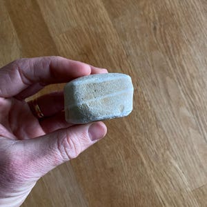May include: A small, rectangular stone with light beige and white horizontal stripes. The stone is held in a hand, showcasing its natural texture and shape. The stone appears to be smooth and worn, suggesting it may be a decorative item or a piece of natural art.