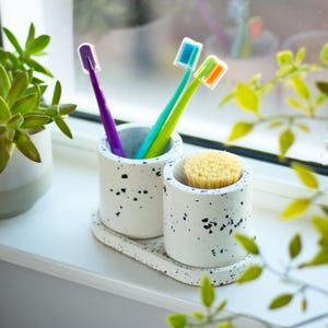 Pode incluir: Dois porta-escovas de dentes de cerâmica branca com manchas pretas. Um porta-escovas tem três escovas de dentes, uma roxa, uma verde e uma laranja. O outro porta-escovas tem uma escova de cerdas naturais. Os porta-escovas estão sobre uma bancada branca.
