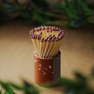 May include: A collection of matches with brown tips, arranged in a decorative, cylindrical holder. The holder is a mix of orange and green, with white speckles and a honeycomb pattern on the side. The matches are a light yellow colour.