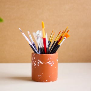 Può includere: Un portapennelli in ceramica color terracotta con macchie bianche, pieno di pennelli da pittura con manici arancioni e neri.