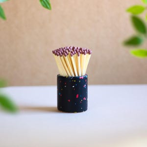 May include: A black ceramic matchstick holder with colourful speckles. The holder is filled with wooden matches with brown tips.