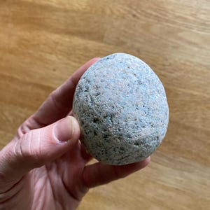 May include: A rounded, grey-blue stone with a speckled surface. The stone is held in a hand, highlighting its natural texture and shape. The stone appears to be about the size of a small apple.
