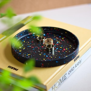 Könnte beinhalten: Ein schwarzes Tablett mit bunten, konfettiartigen Flecken. Das Tablett ist mit goldenen Ringen gefüllt. Das Tablett liegt auf einem Buch mit dem Text "HOME orla kiely PARIS" auf dem Einband.