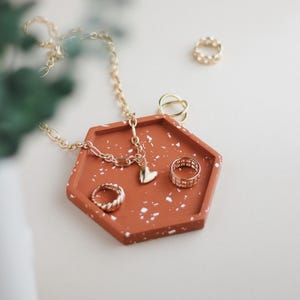 Könnte beinhalten: Eine sechseckige Terrakotta-Schale mit weißen Flecken, die eine goldene Kettenkette mit einem Herz-Anhänger, einen goldenen Ring und einen goldenen Ohrring enthält.