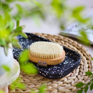 Può includere: Uno spazzolino per il corpo in legno con setole naturali su un vassoio macchiato di bianco e nero. Il vassoio è su una superficie di vimini intrecciato.