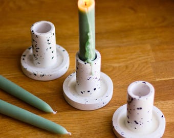 May include: Three white ceramic candle holders with black speckles. The center holder has a green candle with a lit flame. Two additional candles are not lit.