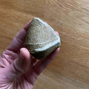 Peut inclure: Une petite pierre triangulaire à la surface rugueuse et texturée. La pierre présente un mélange de couleurs beige, verte et blanche, avec une bande blanche proéminente. La pierre est tenue dans une main, mettant en valeur sa beauté naturelle et sa forme unique.