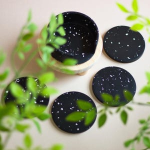 May include: Set of four black and white speckled coasters with a matching coaster holder. The coasters are round and have a speckled design with white flecks on a black background. The coaster holder is a wooden base with a black and white speckled top.