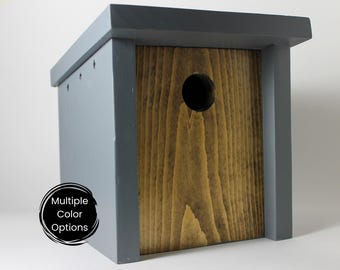 Puede incluir: Una casita de pájaros de madera gris con un panel frontal de madera natural y un agujero de entrada redondo. La casita de pájaros está hecha de madera y tiene un diseño sencillo.