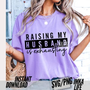 May include: A light purple t-shirt with the text "RAISING MY HUSBAND is exhausting" in black. The shirt is a crew neck style and has short sleeves. The words "INSTANT DOWNLOAD" and "SVG/PNG IMKA LIFE" are also visible.
