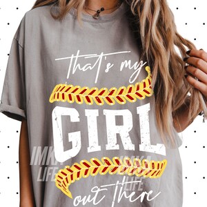 Peut inclure: T-shirt gris avec l'inscription blanche "That's my GIRL out there". Le mot "GIRL" est en grandes lettres grasses. Un motif de softball jaune et rouge est au-dessus et en dessous du mot "GIRL". Le t-shirt a des manches retrouss&eacute;es.