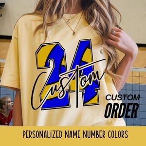 May include: A light yellow t-shirt with a large blue and yellow number 24 design. The word "Custom" is written across the number in a script font. The words "Custom Order" and "Personalized Name Number Colors" are also visible.