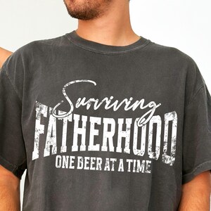 Puede incluir: Una camiseta gris oscura con un gr&aacute;fico blanco desgastado que dice "Surviving Fatherhood One Beer at a Time."